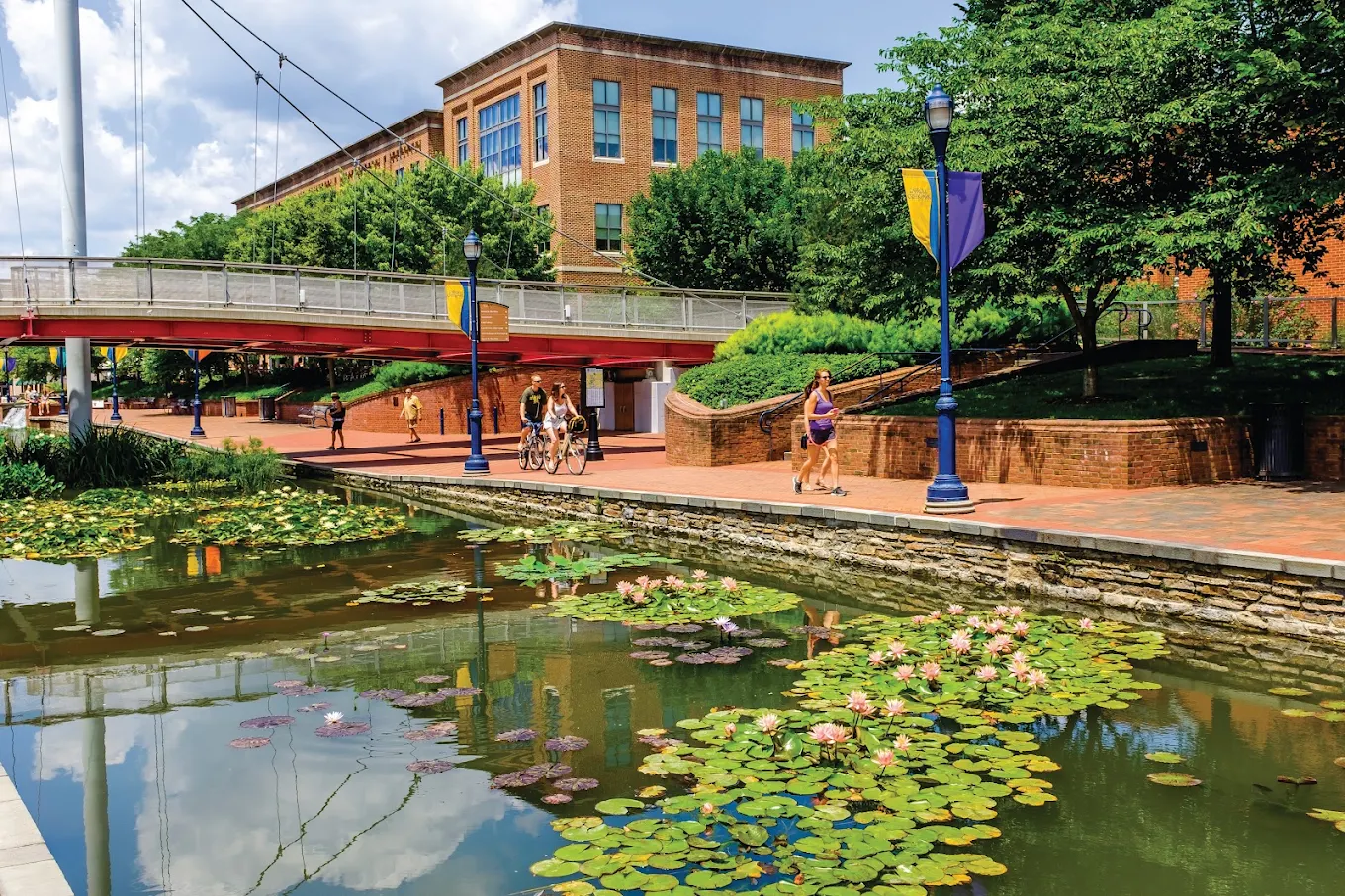 Carroll Creek Park in Downtown Frederick, Maryland, with pedestrians beside the canal, flowering water lilies, a pedestrian bridge and brick buildings.